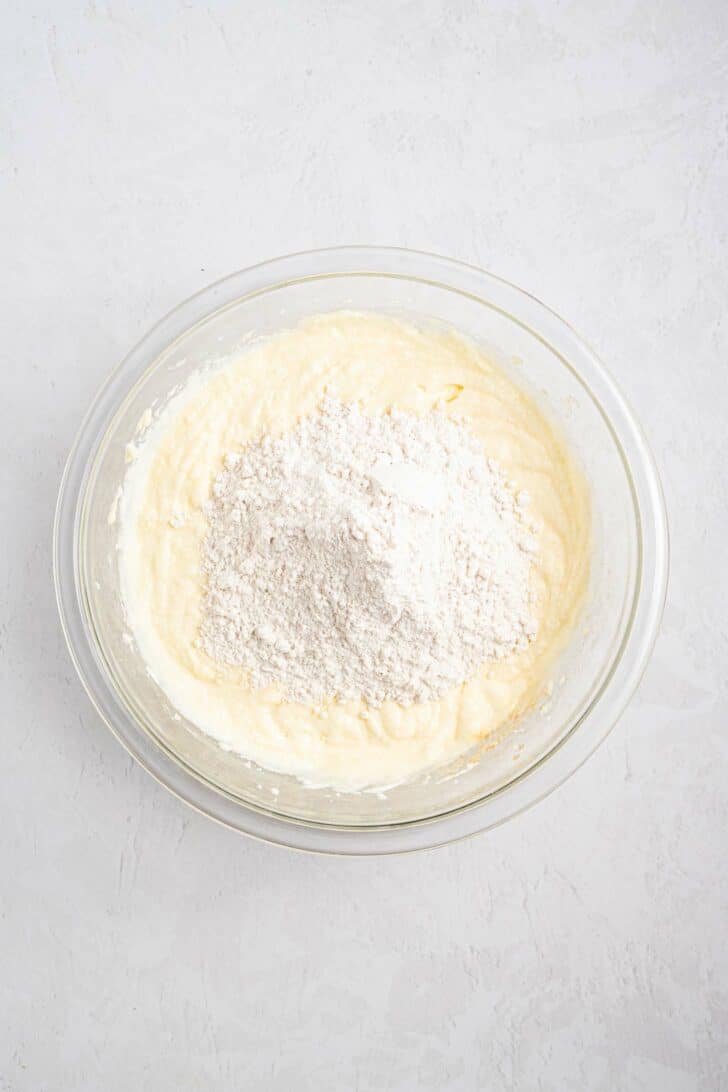 A glass bowl containing a creamy yellow batter with a mound of white flour on top, ready to be mixed, sits on a light textured surface.