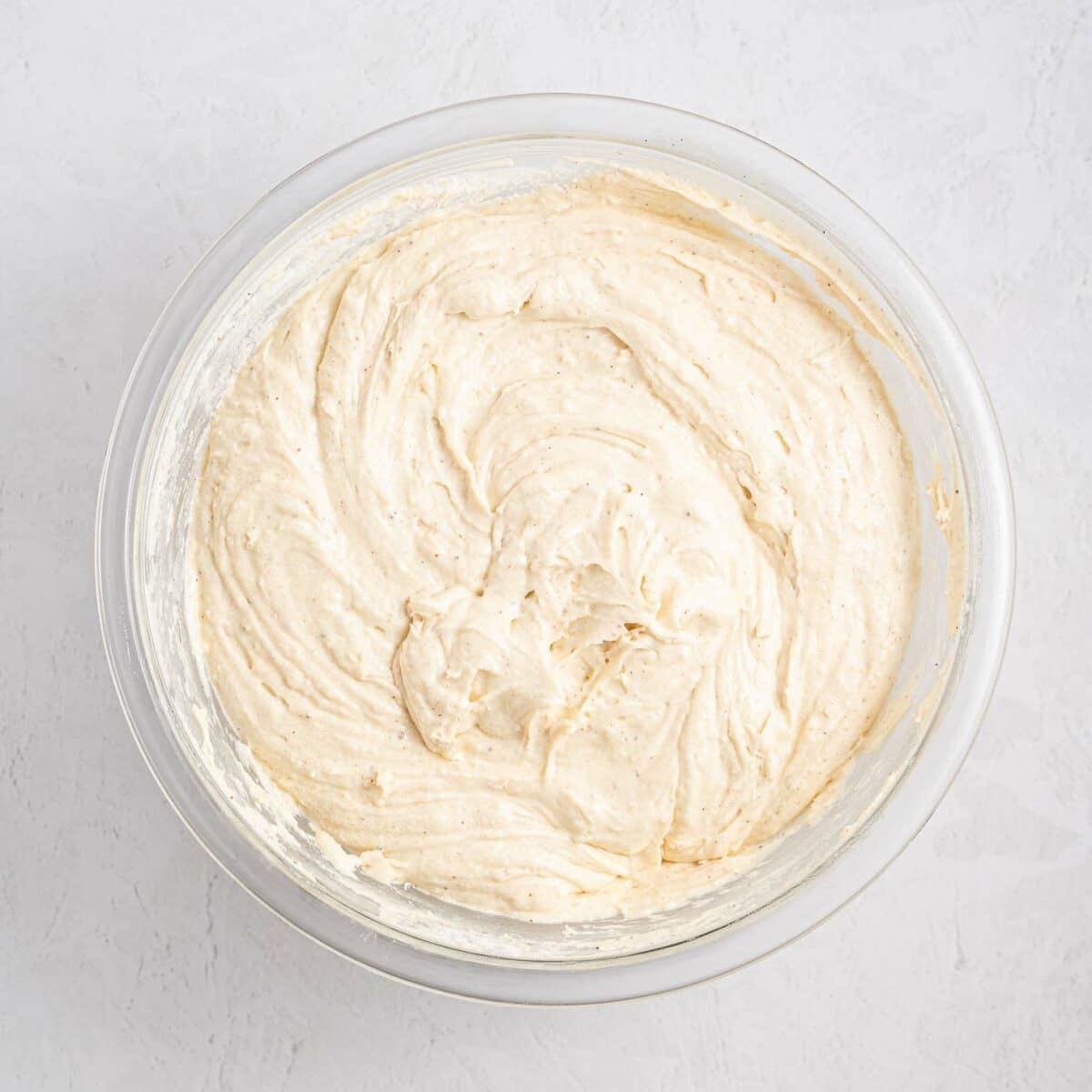 A glass bowl filled with creamy, light beige cake batter, sitting on a white textured surface.