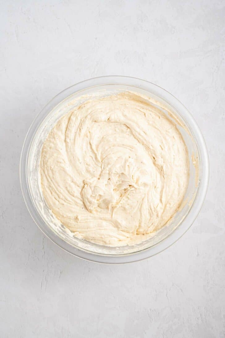 A glass bowl filled with creamy, light beige cake batter, sitting on a white textured surface.
