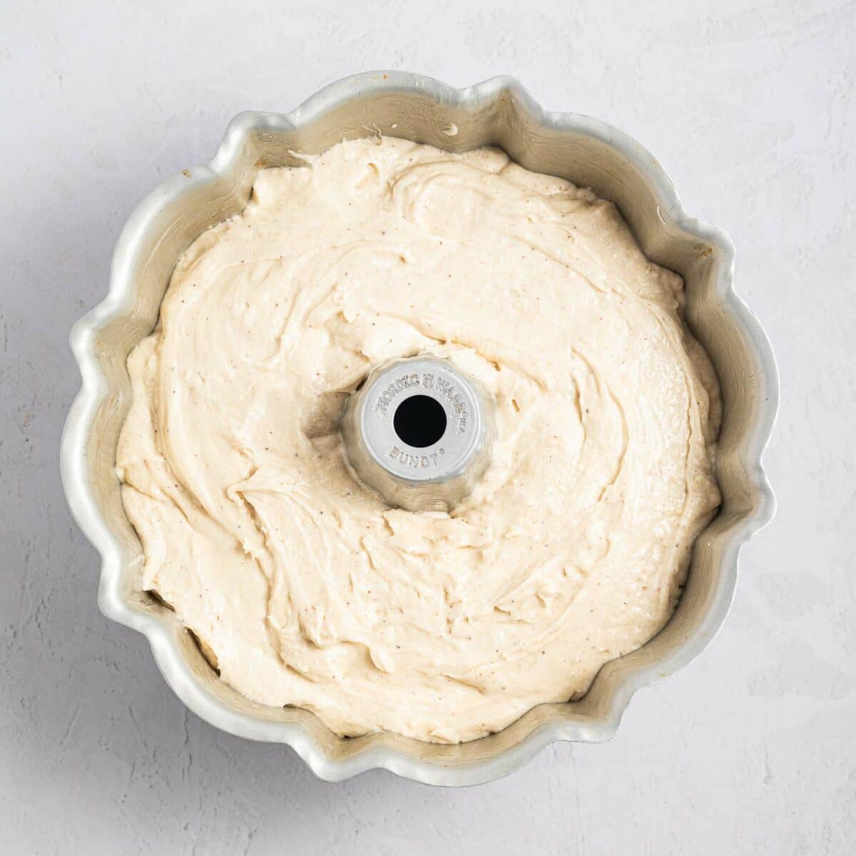 A bundt pan filled with creamy, light-colored cake batter sits on a light, textured surface, ready to be baked.