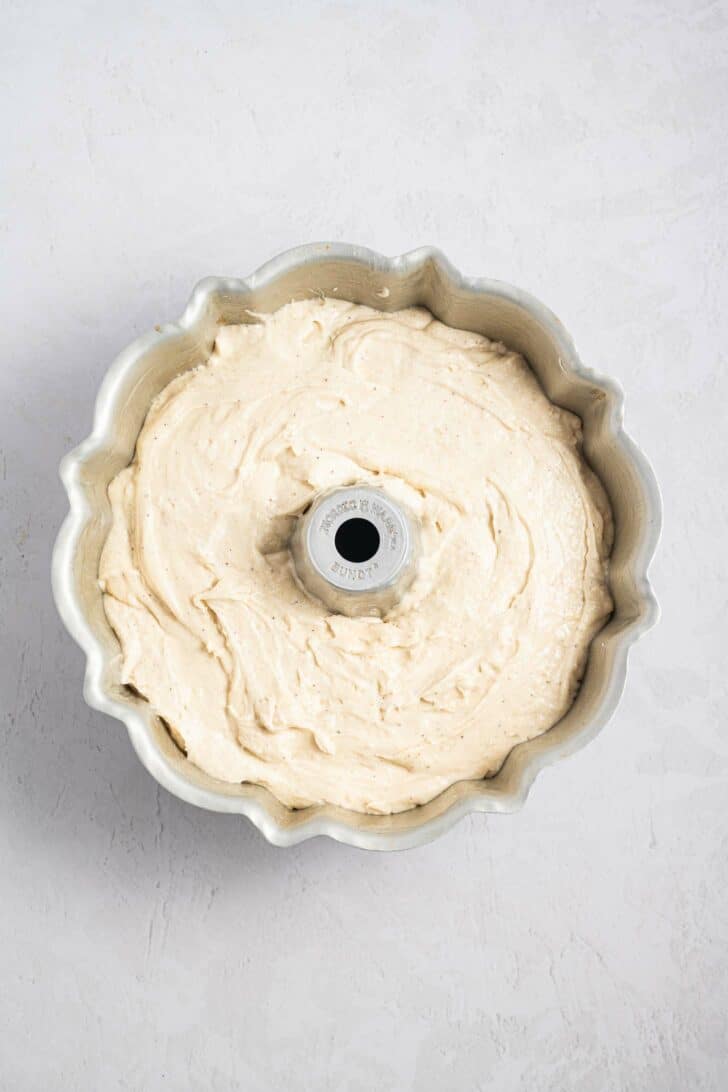 A bundt pan filled with creamy, light-colored cake batter sits on a light, textured surface, ready to be baked.