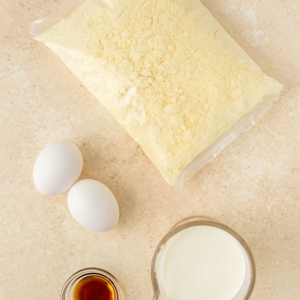A bag of cake mix, two eggs, a small glass cup of vanilla extract, and a measuring cup of milk are arranged on a light-colored countertop.