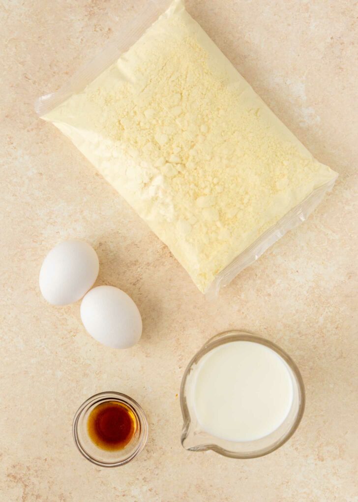 A bag of cake mix, two eggs, a small glass cup of vanilla extract, and a measuring cup of milk are arranged on a light-colored countertop.