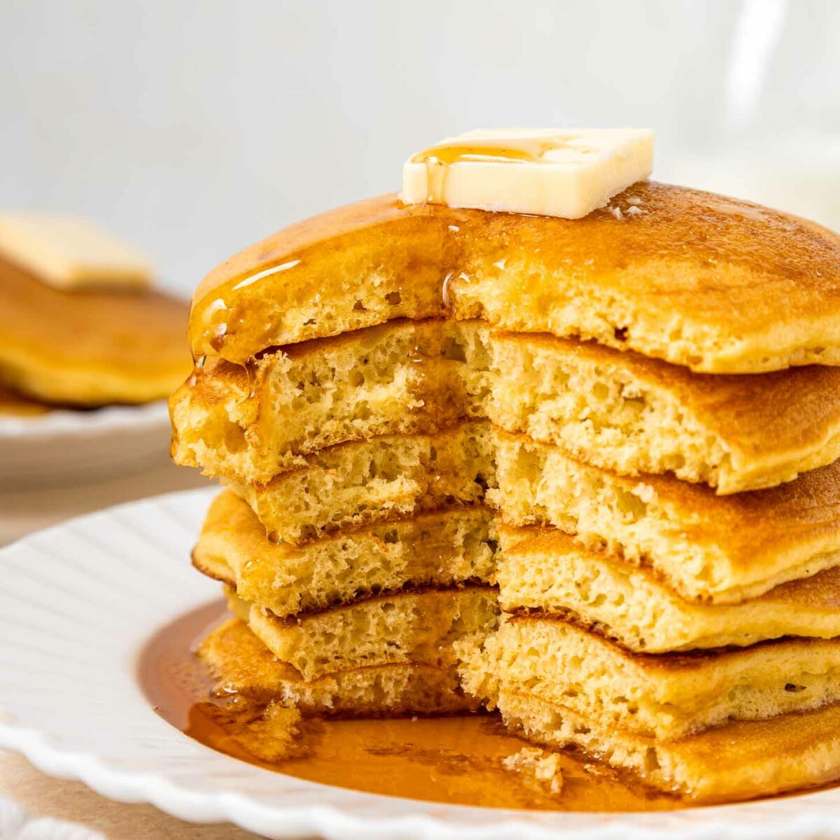 A stack of fluffy pancakes with a pat of butter on top, drizzled with syrup, sits on a white plate. A wedge has been cut from the stack, showing the pancakes’ soft, airy texture.