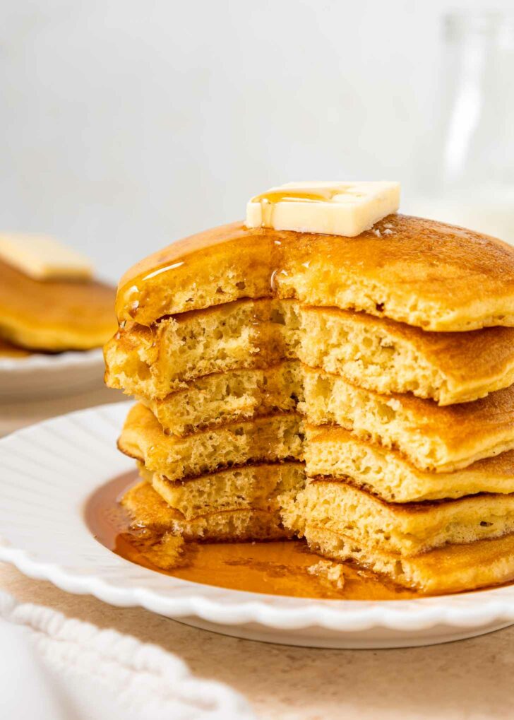 A stack of fluffy pancakes with a pat of butter on top, drizzled with syrup, sits on a white plate. A wedge has been cut from the stack, showing the pancakes’ soft, airy texture.