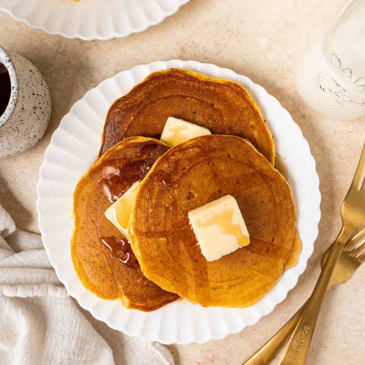 Three pancakes topped with butter and syrup are served on a white plate, with a fork and knife beside it. A small pitcher of syrup, a glass of milk, and a stack of pancakes are nearby.