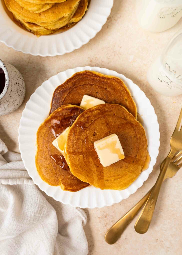 Three pancakes topped with butter and syrup are served on a white plate, with a fork and knife beside it. A small pitcher of syrup, a glass of milk, and a stack of pancakes are nearby.