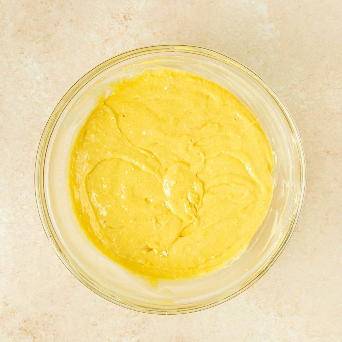 A clear glass bowl filled with yellow cake batter sits on a light beige countertop.