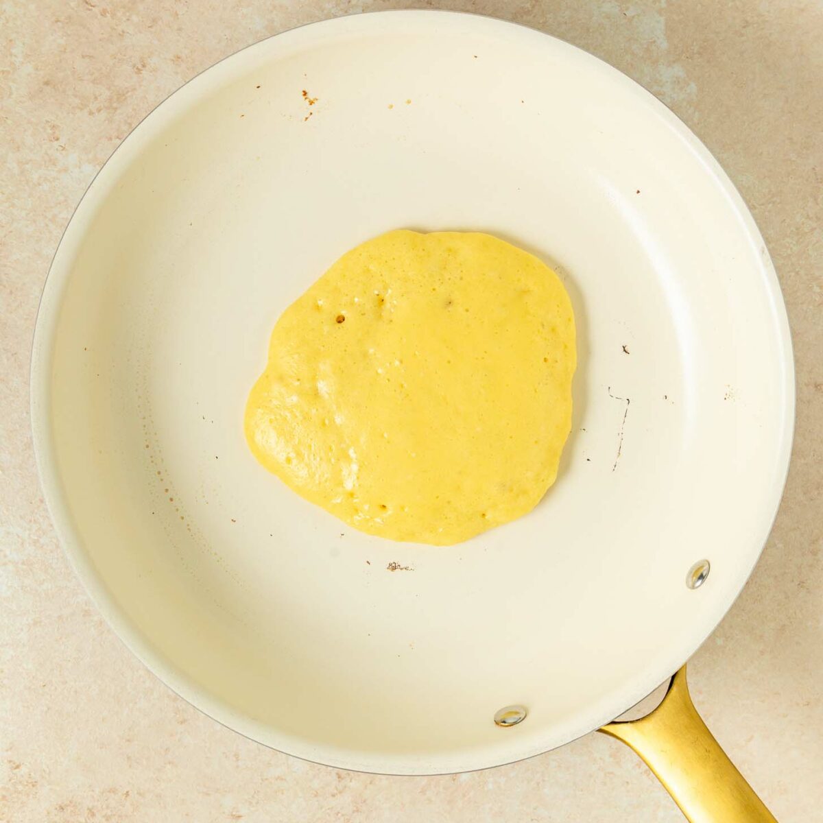 A single pancake cooking in a white nonstick skillet with a gold handle, resting on a light beige countertop.