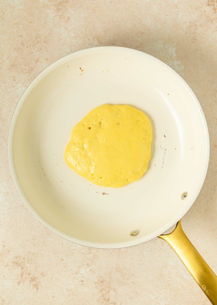 A single pancake cooking in a white nonstick skillet with a gold handle, resting on a light beige countertop.