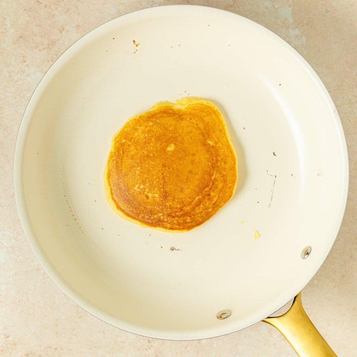 A single golden-brown pancake cooking in a white nonstick skillet with a gold handle, viewed from above. The pan sits on a light-colored surface.
