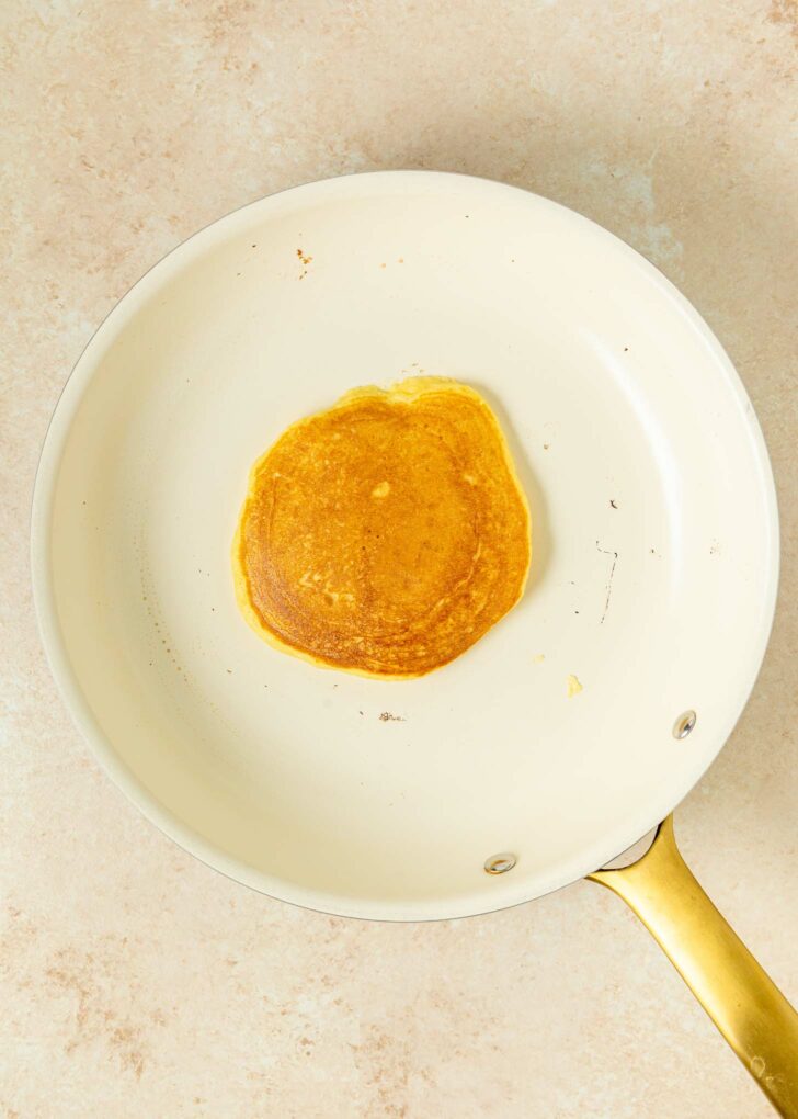 A single golden-brown pancake cooking in a white nonstick skillet with a gold handle, viewed from above. The pan sits on a light-colored surface.