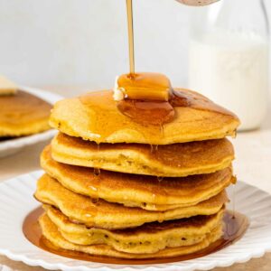 A stack of fluffy pancakes topped with a pat of butter, with maple syrup being poured over them, on a white plate with a bottle of milk in the background.