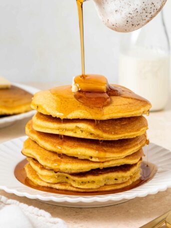 A stack of fluffy pancakes topped with a pat of butter, with maple syrup being poured over them, on a white plate with a bottle of milk in the background.