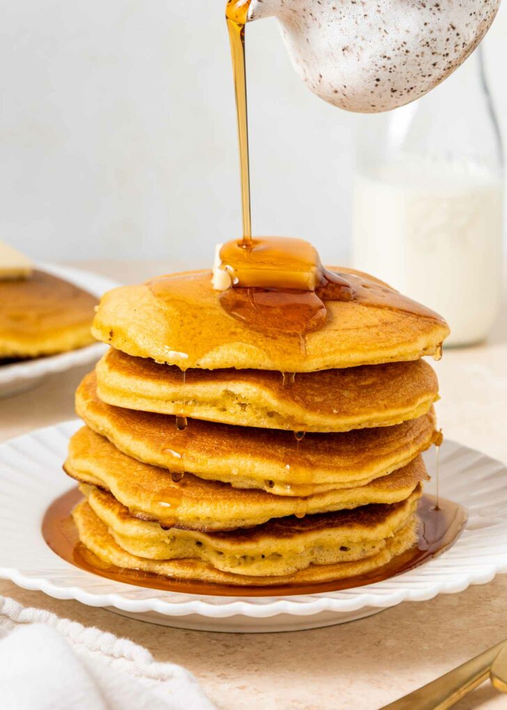 A stack of fluffy pancakes topped with a pat of butter, with maple syrup being poured over them, on a white plate with a bottle of milk in the background.