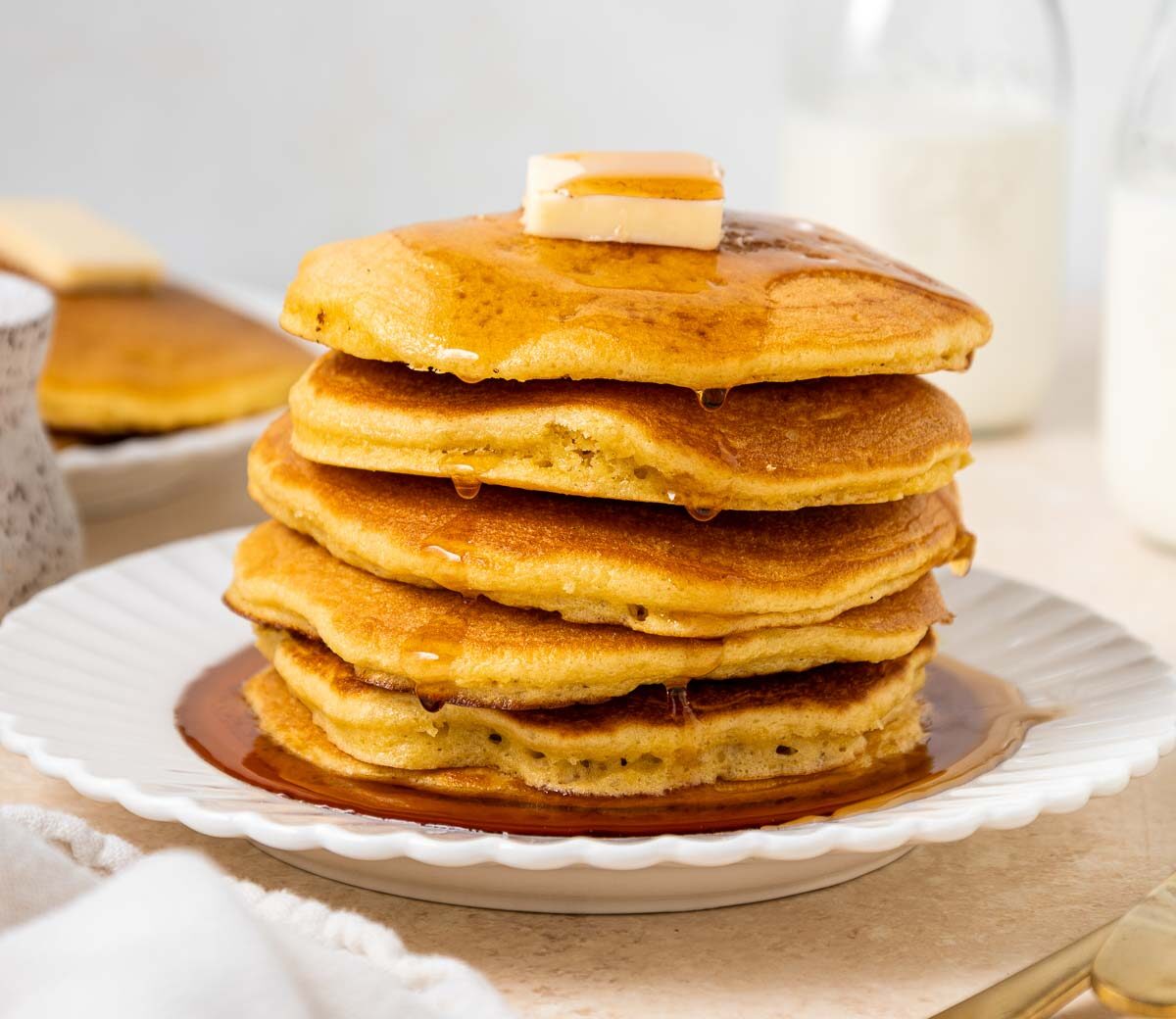 A stack of golden pancakes topped with butter and syrup sits on a white plate. In the background are bottles of milk, a speckled mug, a fork, a knife, and another plate of pancakes.
