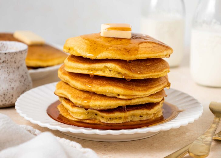 A stack of golden pancakes topped with butter and syrup sits on a white plate. In the background are bottles of milk, a speckled mug, a fork, a knife, and another plate of pancakes.