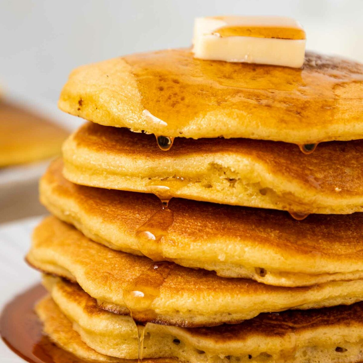 A close-up of a stack of fluffy pancakes topped with a pat of butter and drizzled with syrup, sitting on a white plate. Syrup flows down the sides of the golden-brown pancakes.