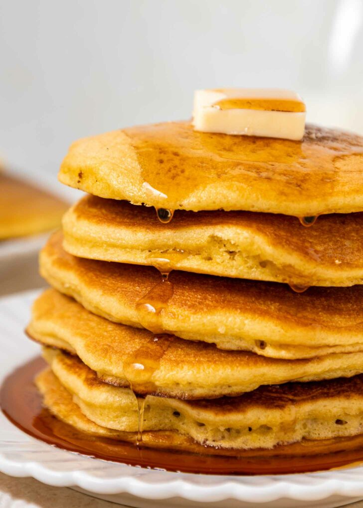 A close-up of a stack of fluffy pancakes topped with a pat of butter and drizzled with syrup, sitting on a white plate. Syrup flows down the sides of the golden-brown pancakes.