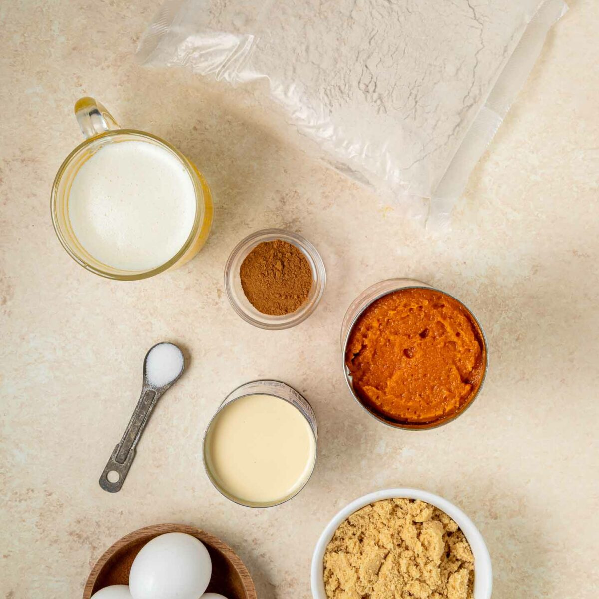 A flat lay of baking ingredients: a bag of cake mix, sweetened condensed milk, a bowl of brown sugar, three eggs, a small bowl of melted butter, a jar of cinnamon, a can of pumpkin puree, and a spoon with salt.