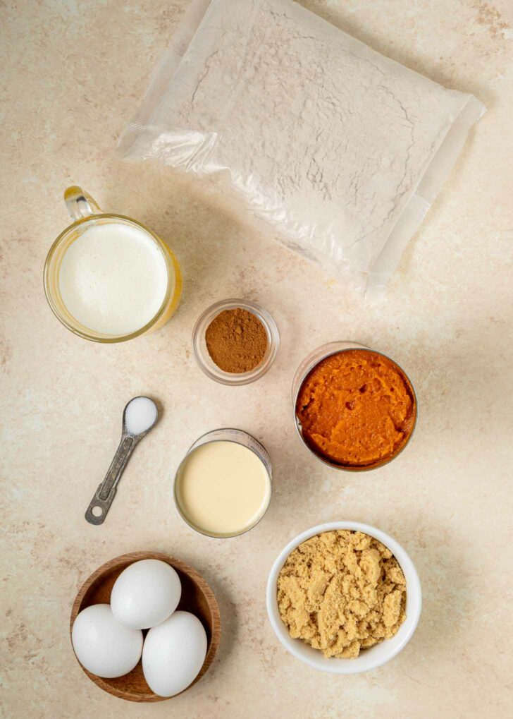 A flat lay of baking ingredients: a bag of cake mix, sweetened condensed milk, a bowl of brown sugar, three eggs, a small bowl of melted butter, a jar of cinnamon, a can of pumpkin puree, and a spoon with salt.