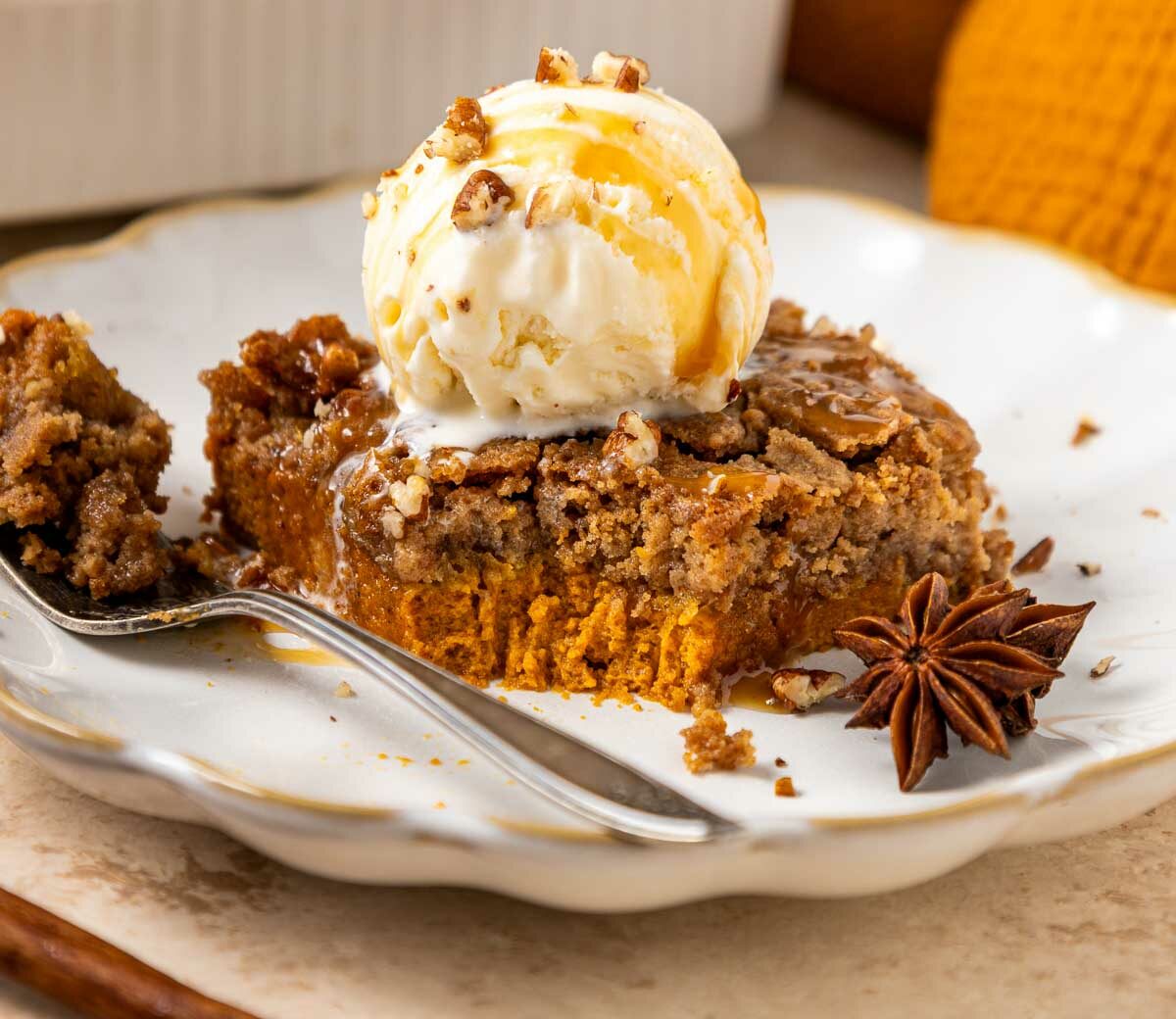 A slice of pumpkin dump cake topped with a scoop of vanilla ice cream, drizzled with caramel sauce, and garnished with chopped nuts and a star anise on a decorative white plate. A fork holds a bite of the cake.