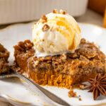 A slice of pumpkin dump cake topped with a scoop of vanilla ice cream, drizzled with caramel sauce and sprinkled with chopped nuts, sits on a plate with a fork and a star anise as garnish.