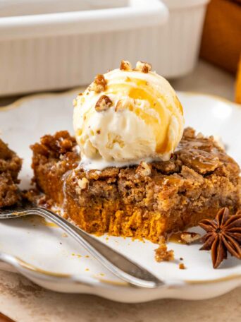 A slice of pumpkin dump cake topped with a scoop of vanilla ice cream, drizzled with caramel sauce and sprinkled with chopped nuts, sits on a plate with a fork and a star anise as garnish.