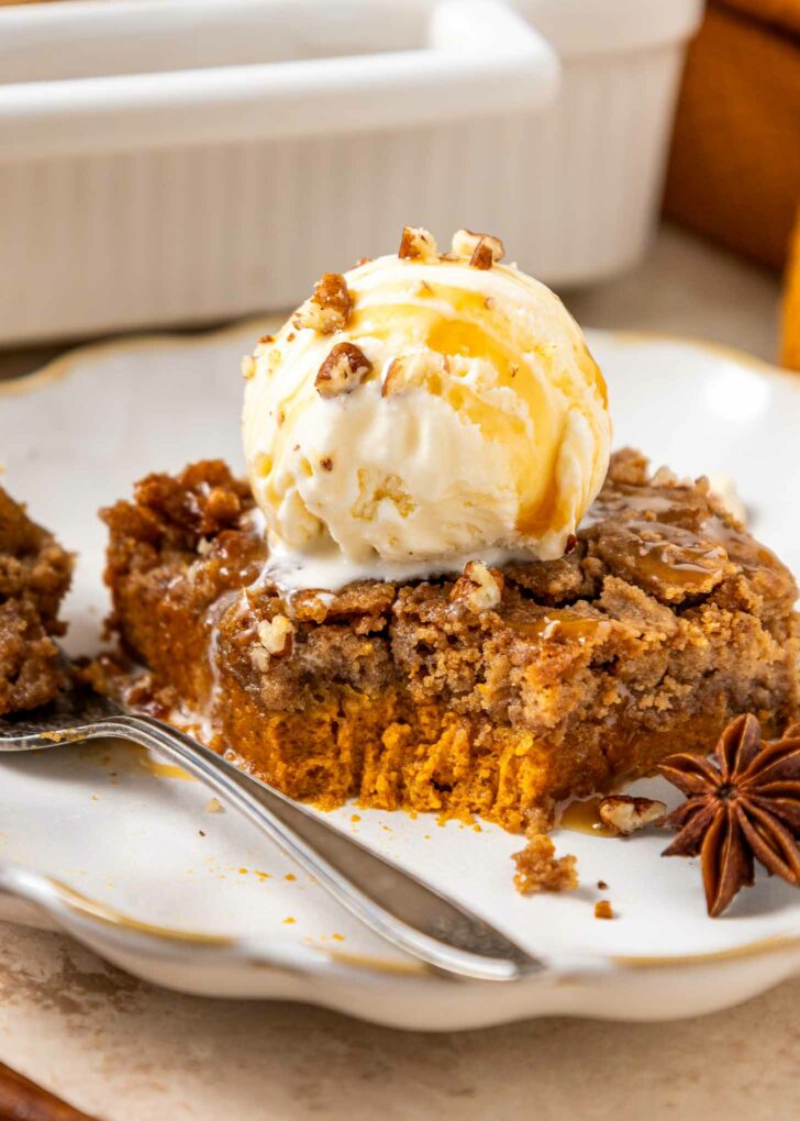 A slice of pumpkin dump cake topped with a scoop of vanilla ice cream, drizzled with caramel sauce and sprinkled with chopped nuts, sits on a plate with a fork and a star anise as garnish.