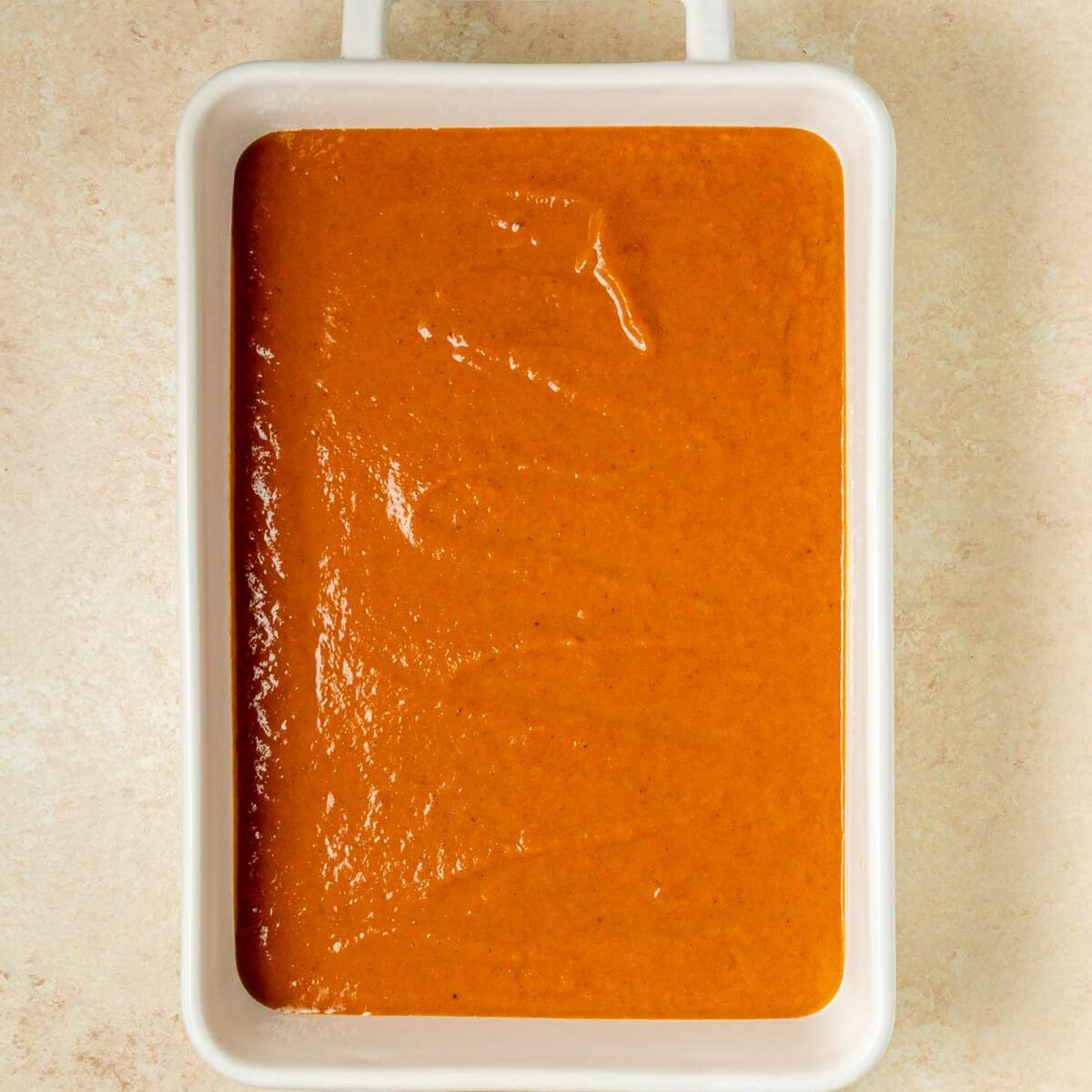 A white rectangular baking dish filled with a smooth layer of orange-brown dump cake batter, set on a light beige textured surface.