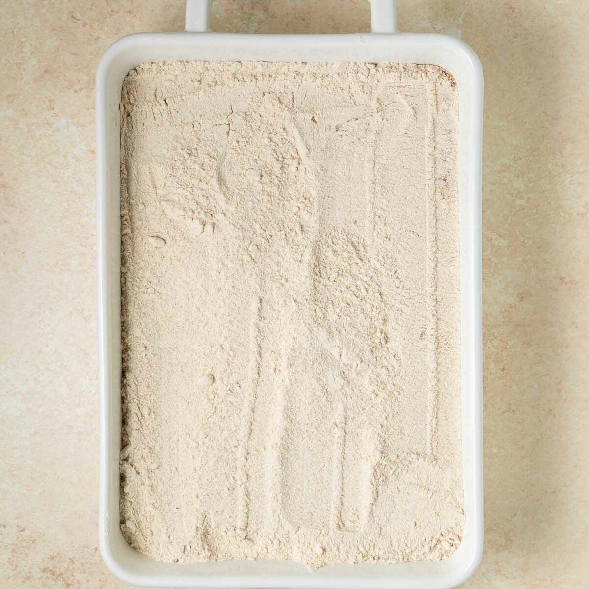 A white rectangular baking dish topped with cake mix, with a lightly smoothed surface, sits on a beige countertop.