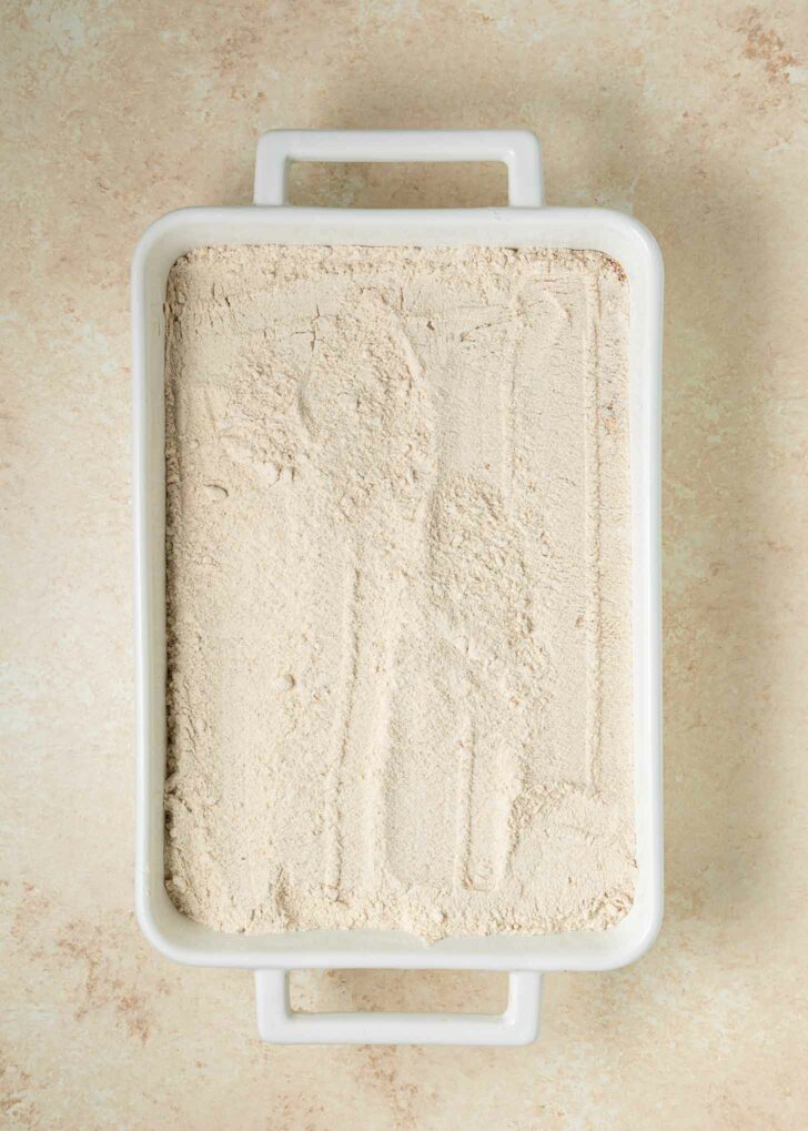 A white rectangular baking dish topped with cake mix, with a lightly smoothed surface, sits on a beige countertop.