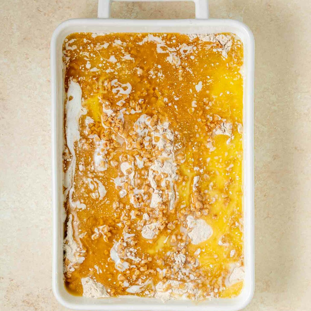 A rectangular white baking dish filled with an unbaked mixture of yellow melted butter, cake mix, and some dry ingredients, showing a marbled texture, on a light-colored countertop.