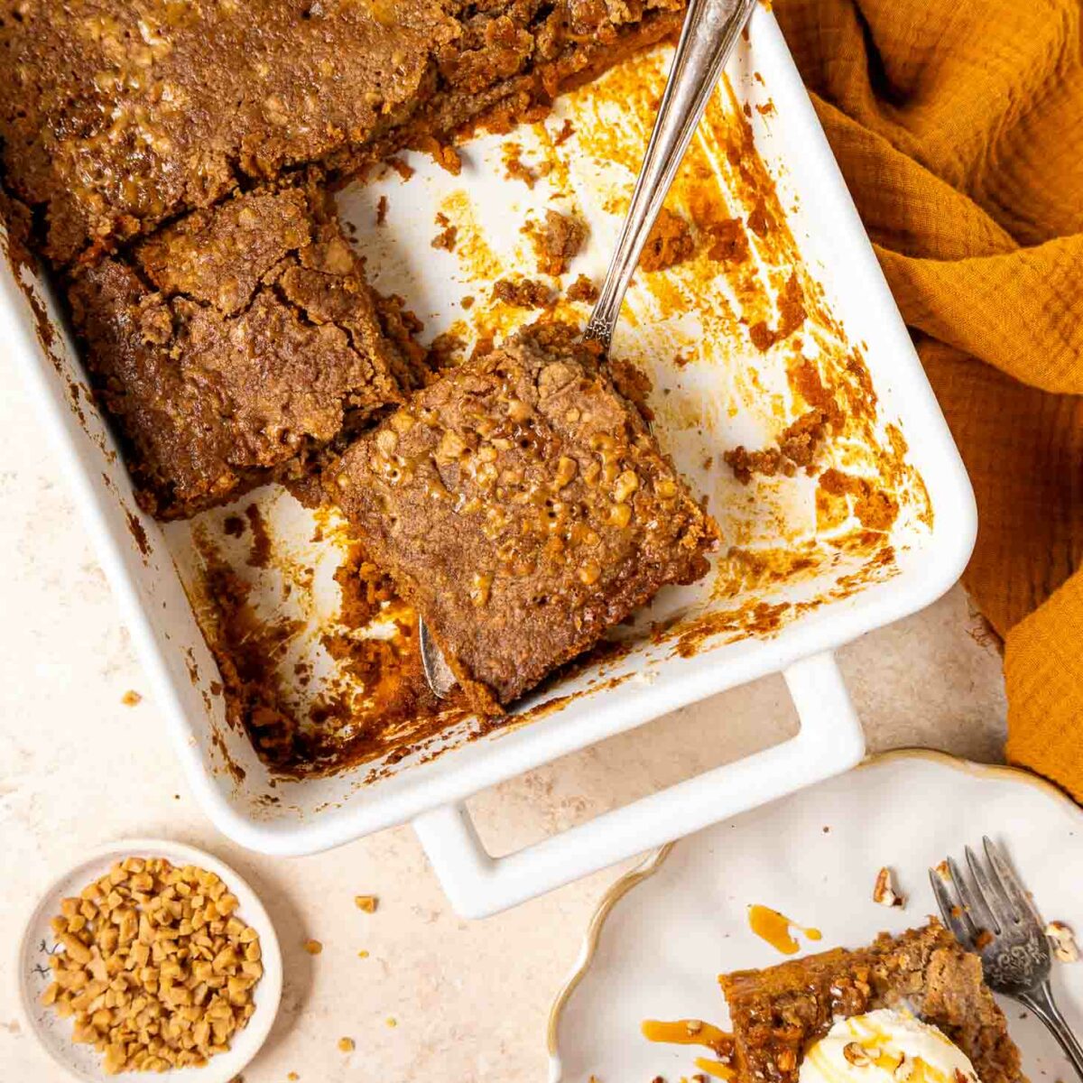 A white baking dish with pumpkin dump cake, one square piece being lifted out with a cake server. A plated slice with ice cream and caramel sauce sits nearby, with a bowl of chopped nuts and a cinnamon stick on the table.