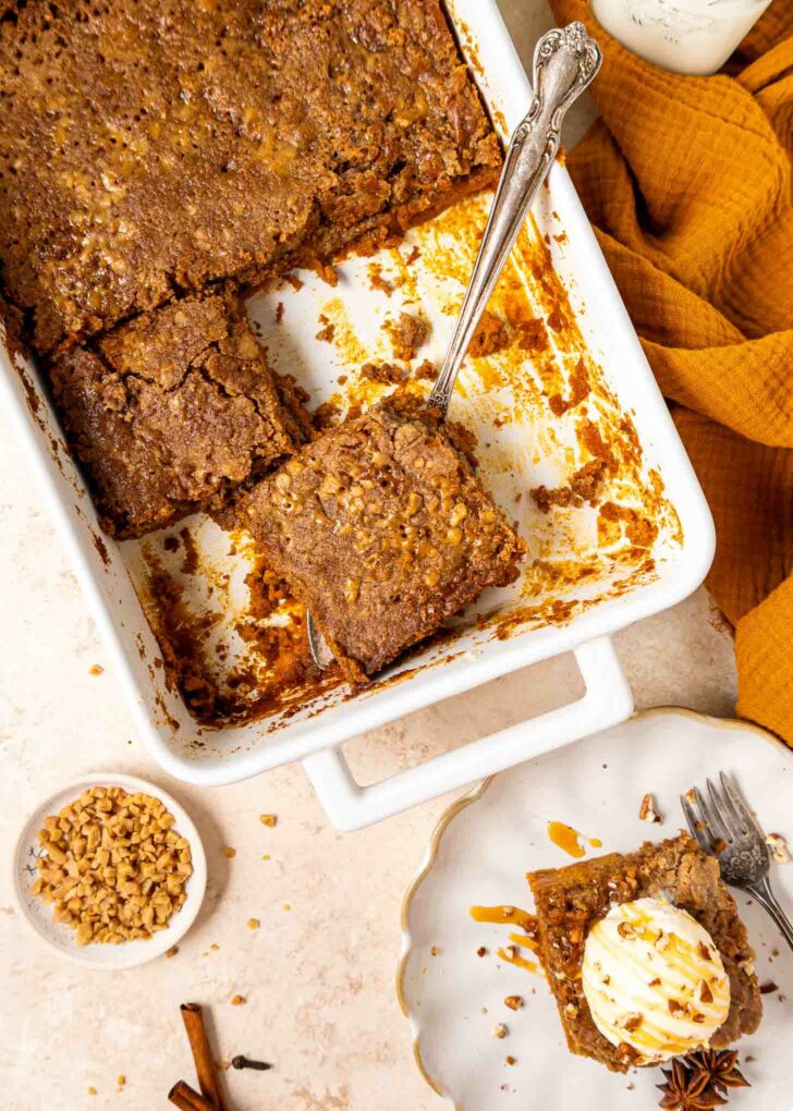 A white baking dish with pumpkin dump cake, one square piece being lifted out with a cake server. A plated slice with ice cream and caramel sauce sits nearby, with a bowl of chopped nuts and a cinnamon stick on the table.