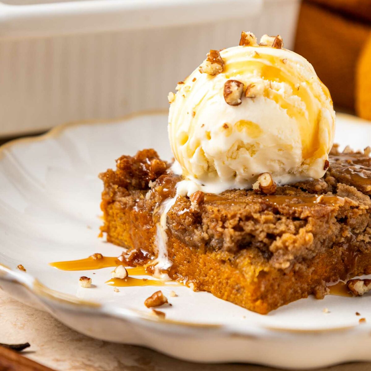 A slice of pumpkin dump cake topped with a scoop of vanilla ice cream, toffee bits, and caramel drizzle on a white plate. A pan of more dessert and a cinnamon stick are in the background.