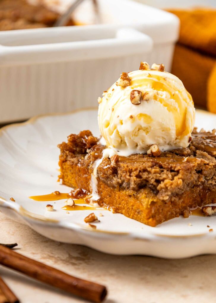A slice of pumpkin dump cake topped with a scoop of vanilla ice cream, toffee bits, and caramel drizzle on a white plate. A pan of more dessert and a cinnamon stick are in the background.