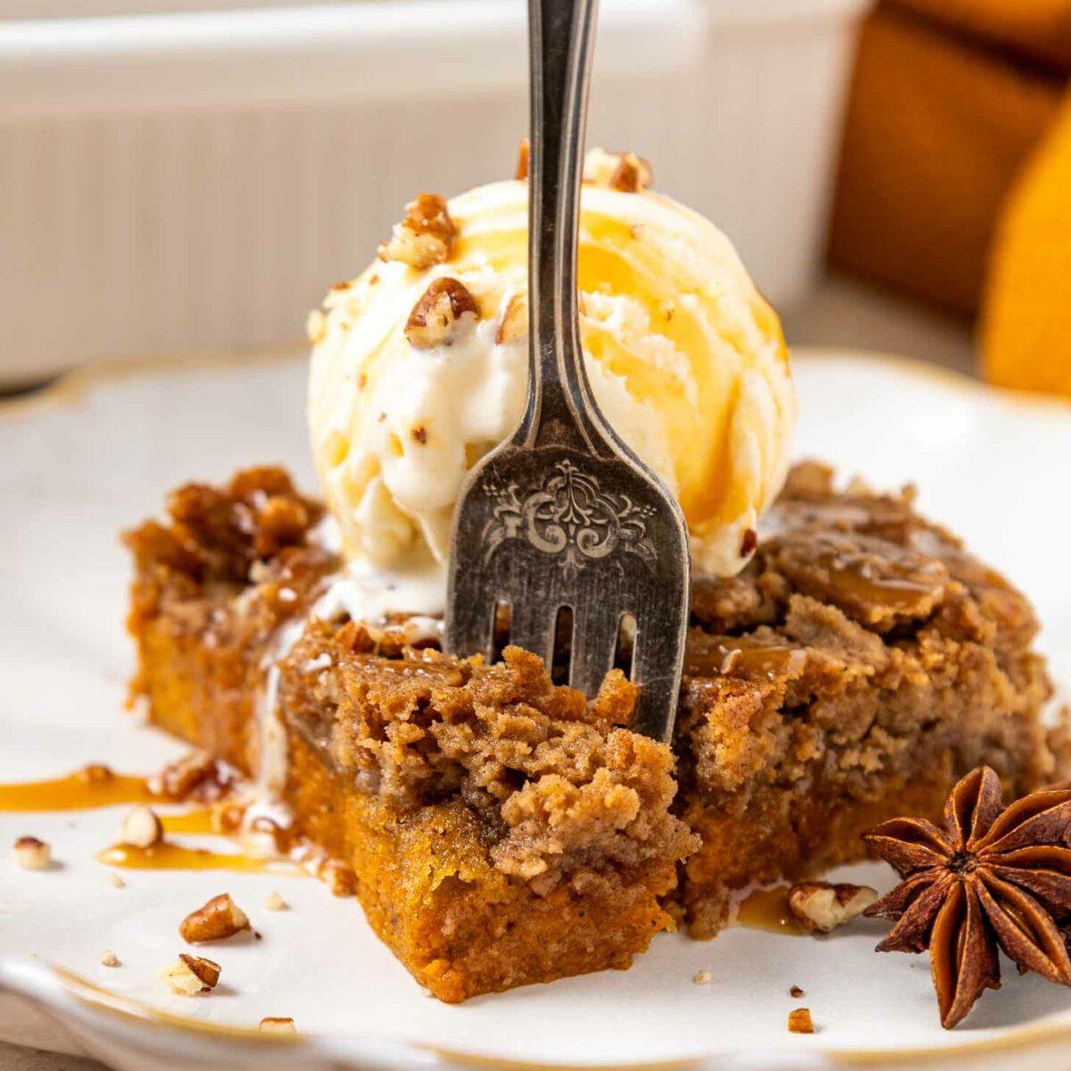 A fork rests in a slice of spiced pumpkin dump cake topped with a scoop of vanilla ice cream, chopped pecans, and caramel drizzle on a white plate. An anise star garnish sits beside the dessert.
