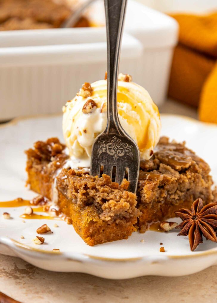 A fork rests in a slice of spiced pumpkin dump cake topped with a scoop of vanilla ice cream, chopped pecans, and caramel drizzle on a white plate. An anise star garnish sits beside the dessert.