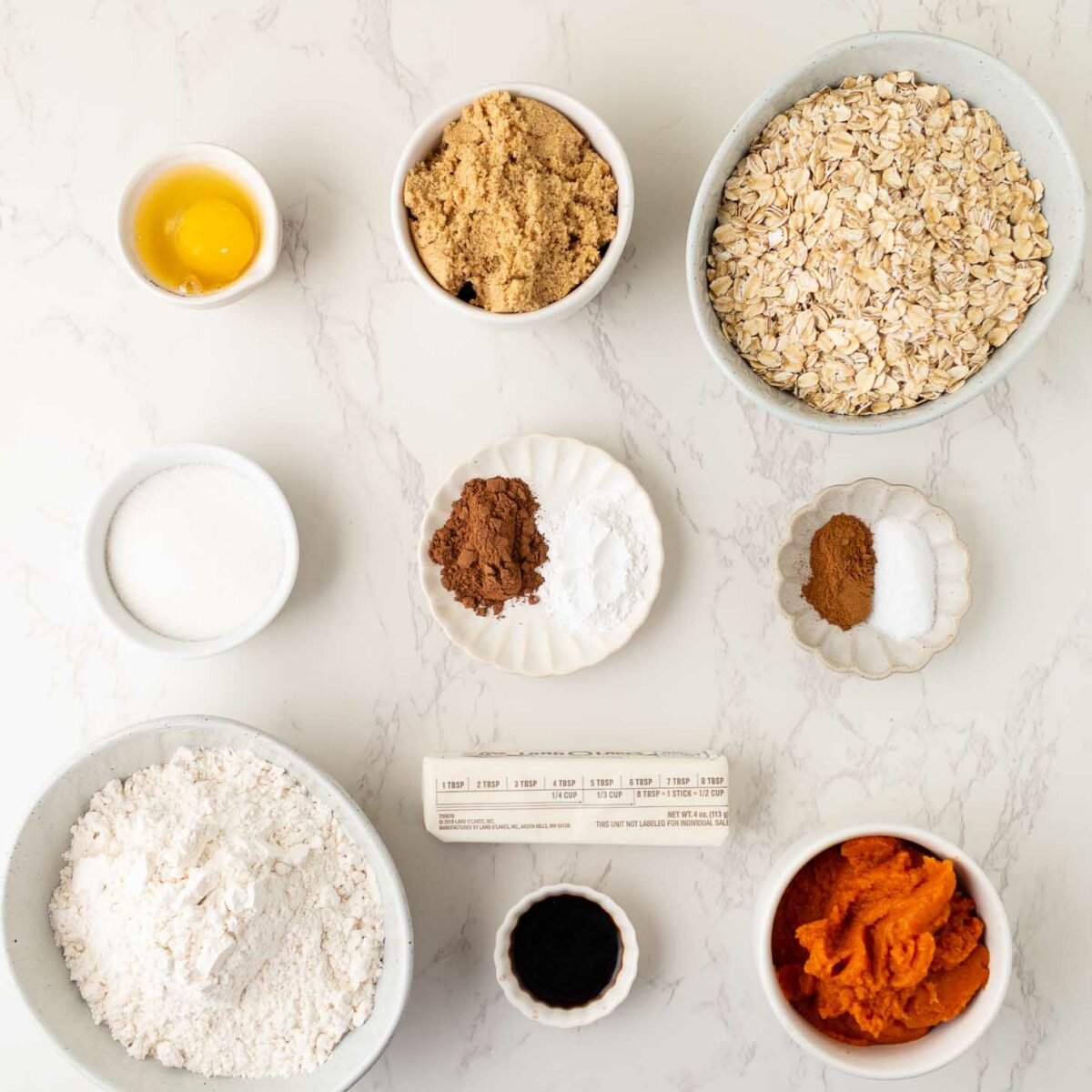 Top-down view of baking ingredients in bowls and plates on a marble surface, including an egg, brown sugar, oats, flour, pumpkin puree, spices, vanilla extract, butter, and a small bowl of milk.