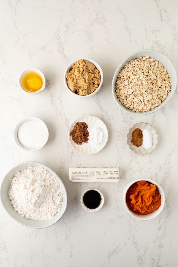 Top-down view of baking ingredients in bowls and plates on a marble surface, including an egg, brown sugar, oats, flour, pumpkin puree, spices, vanilla extract, butter, and a small bowl of milk.