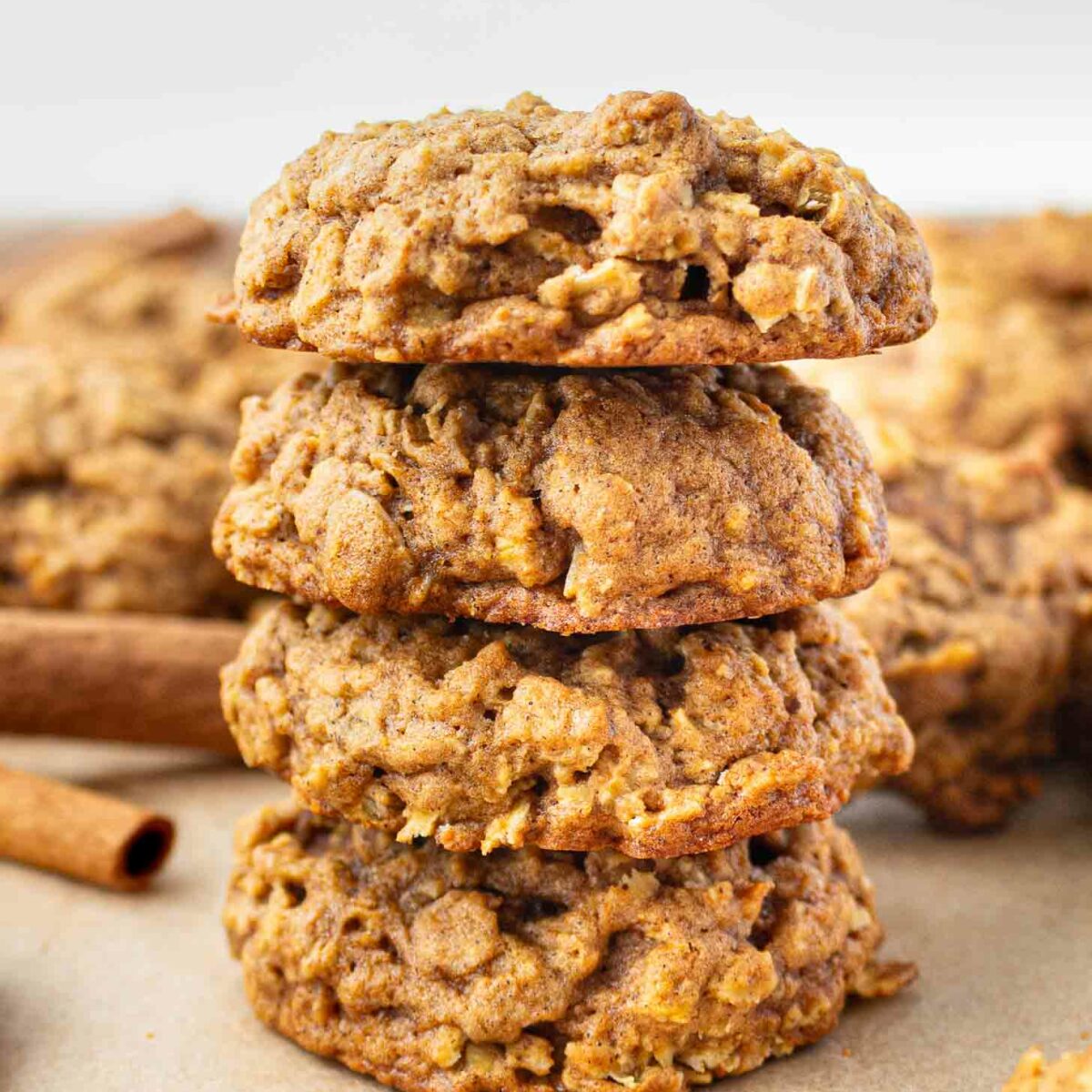 A stack of four pumpkin oatmeal cookies sits on a light surface with more cookies and cinnamon sticks in the background. The cookies have a soft, chunky texture and a golden-brown color.