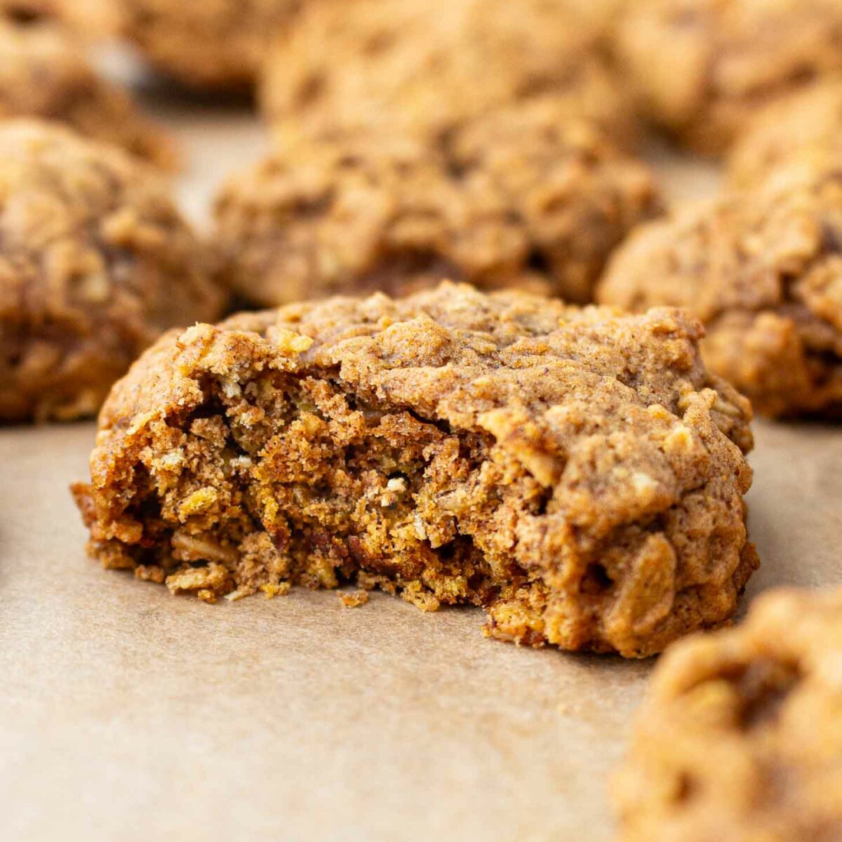 A close-up of a soft, chewy pumpkin oatmeal cookie with a bite taken out, showing its textured inside. More cookies are blurred in the background, all resting on parchment paper.