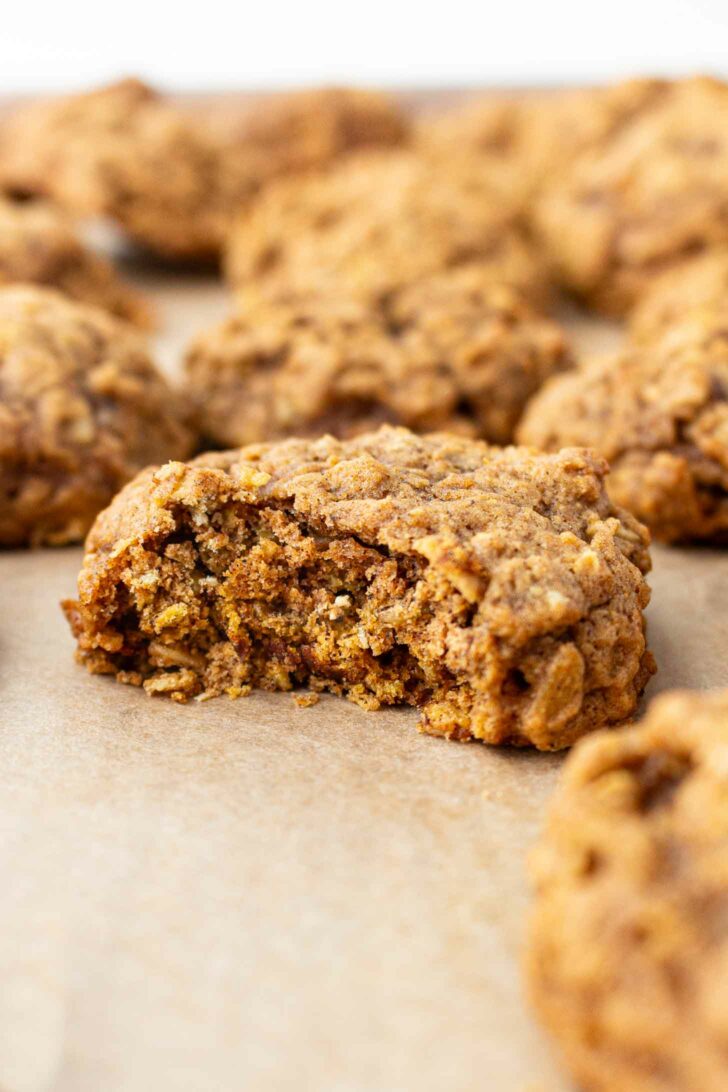 A close-up of a soft, chewy pumpkin oatmeal cookie with a bite taken out, showing its textured inside. More cookies are blurred in the background, all resting on parchment paper.
