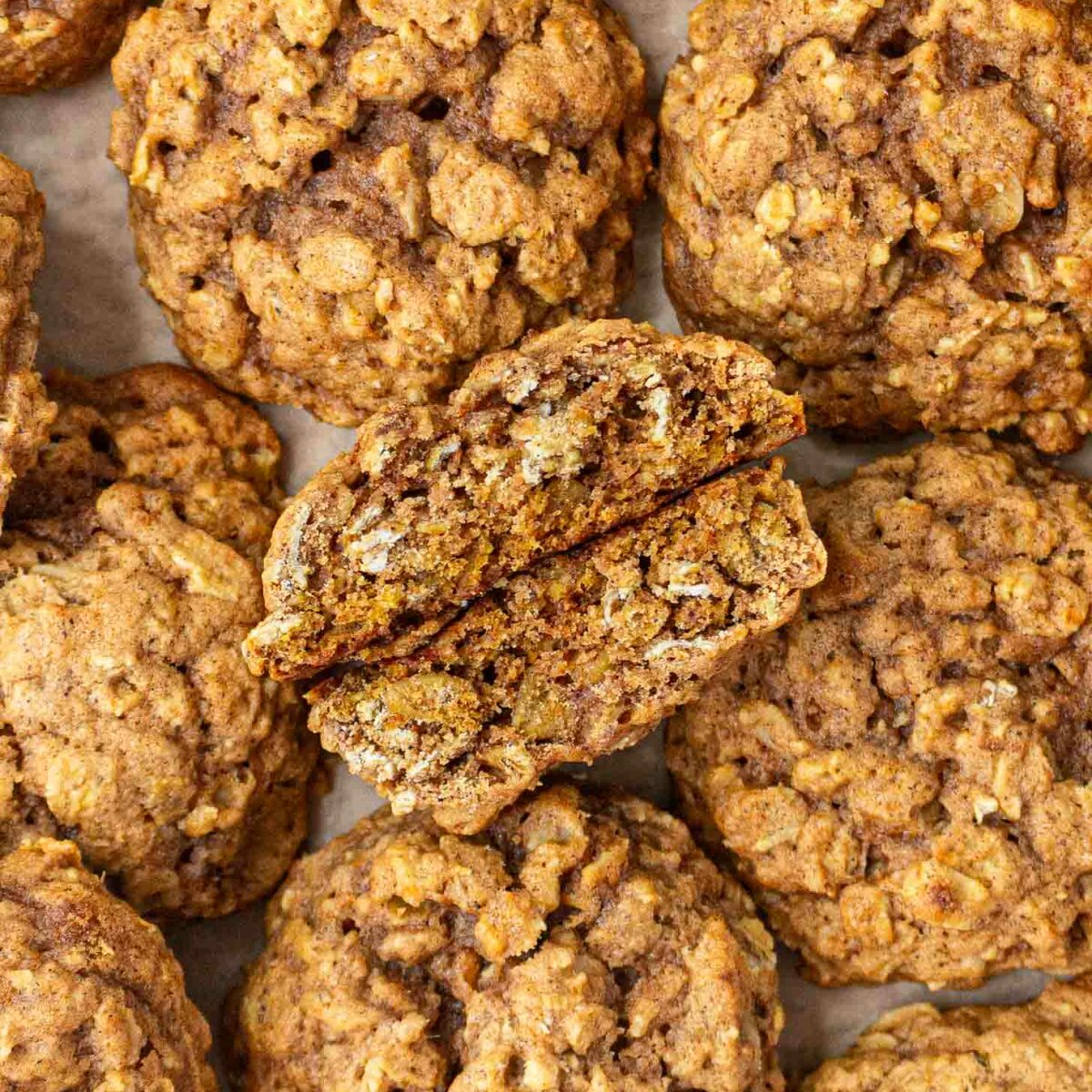 A close-up view of several pumpkin oatmeal cookies on parchment paper, with one cookie broken in half to show its chewy, textured interior.
