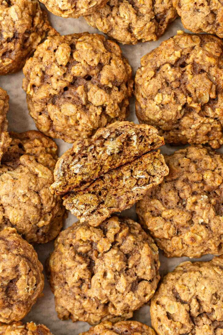 A close-up view of several pumpkin oatmeal cookies on parchment paper, with one cookie broken in half to show its chewy, textured interior.
