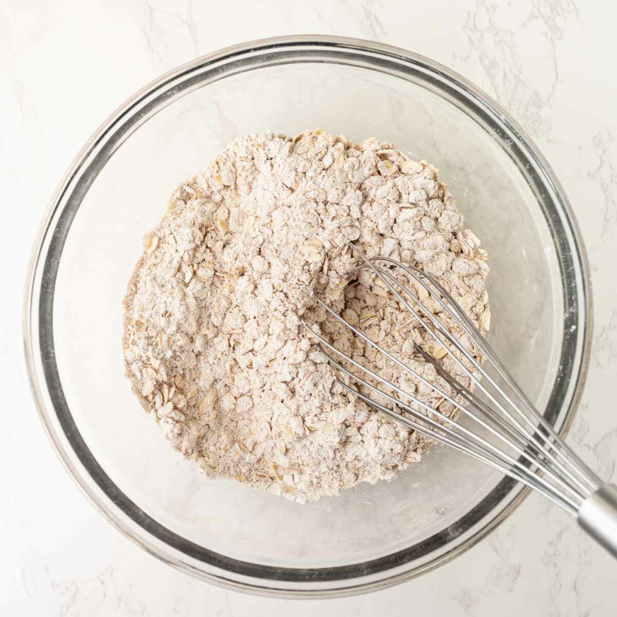 A glass bowl containing a mixture of flour and oats being whisked with a metal whisk on a light-colored marble surface.