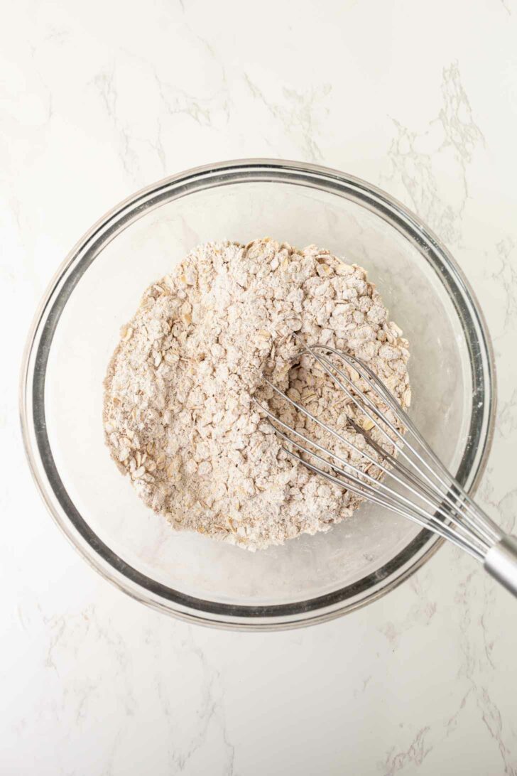 A glass bowl containing a mixture of flour and oats being whisked with a metal whisk on a light-colored marble surface.