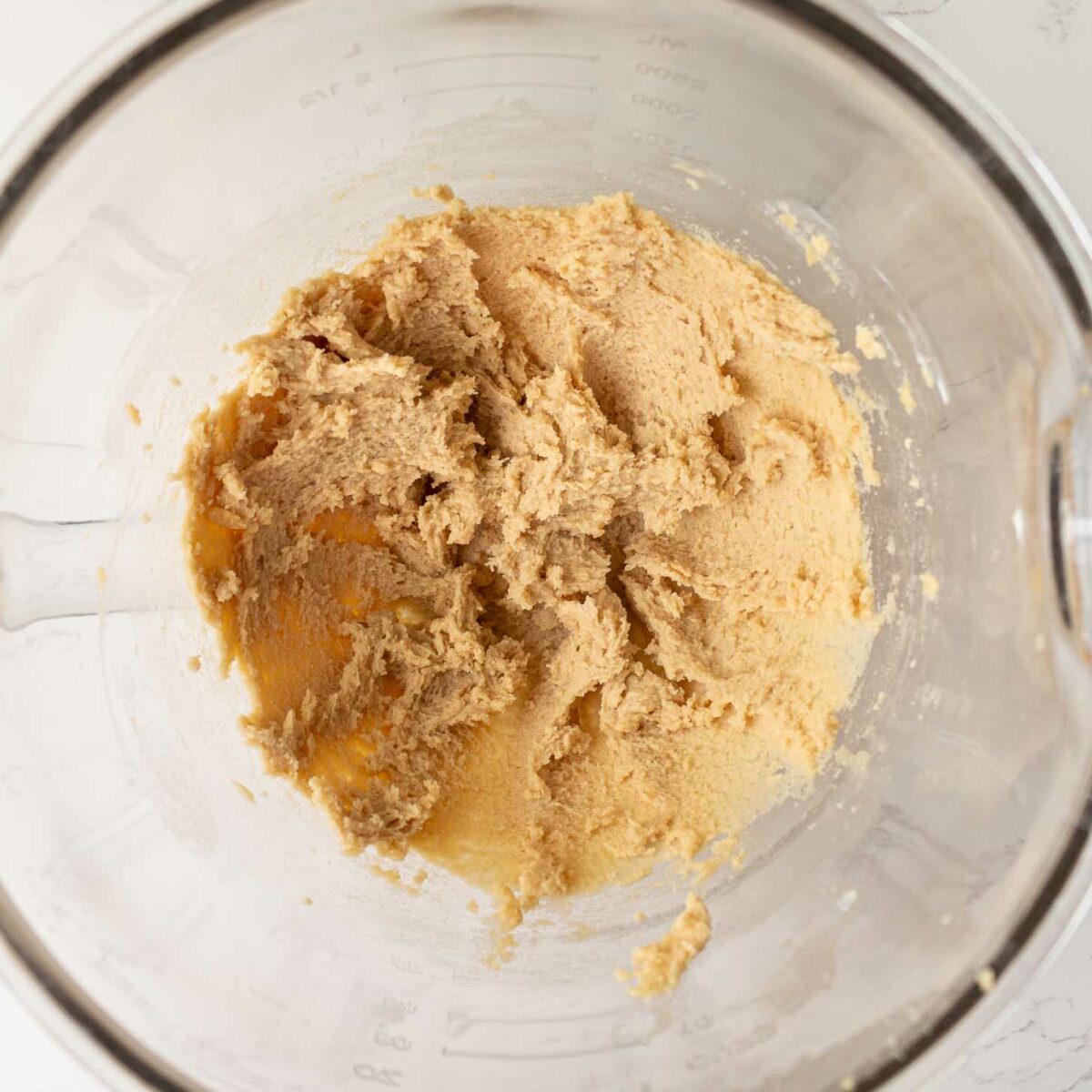 A glass mixing bowl containing light brown cookie dough mixture on a white marble surface, viewed from above.