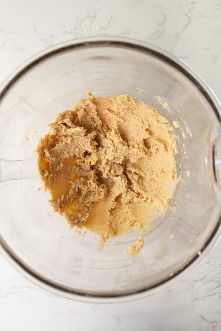 A glass mixing bowl containing light brown cookie dough mixture on a white marble surface, viewed from above.